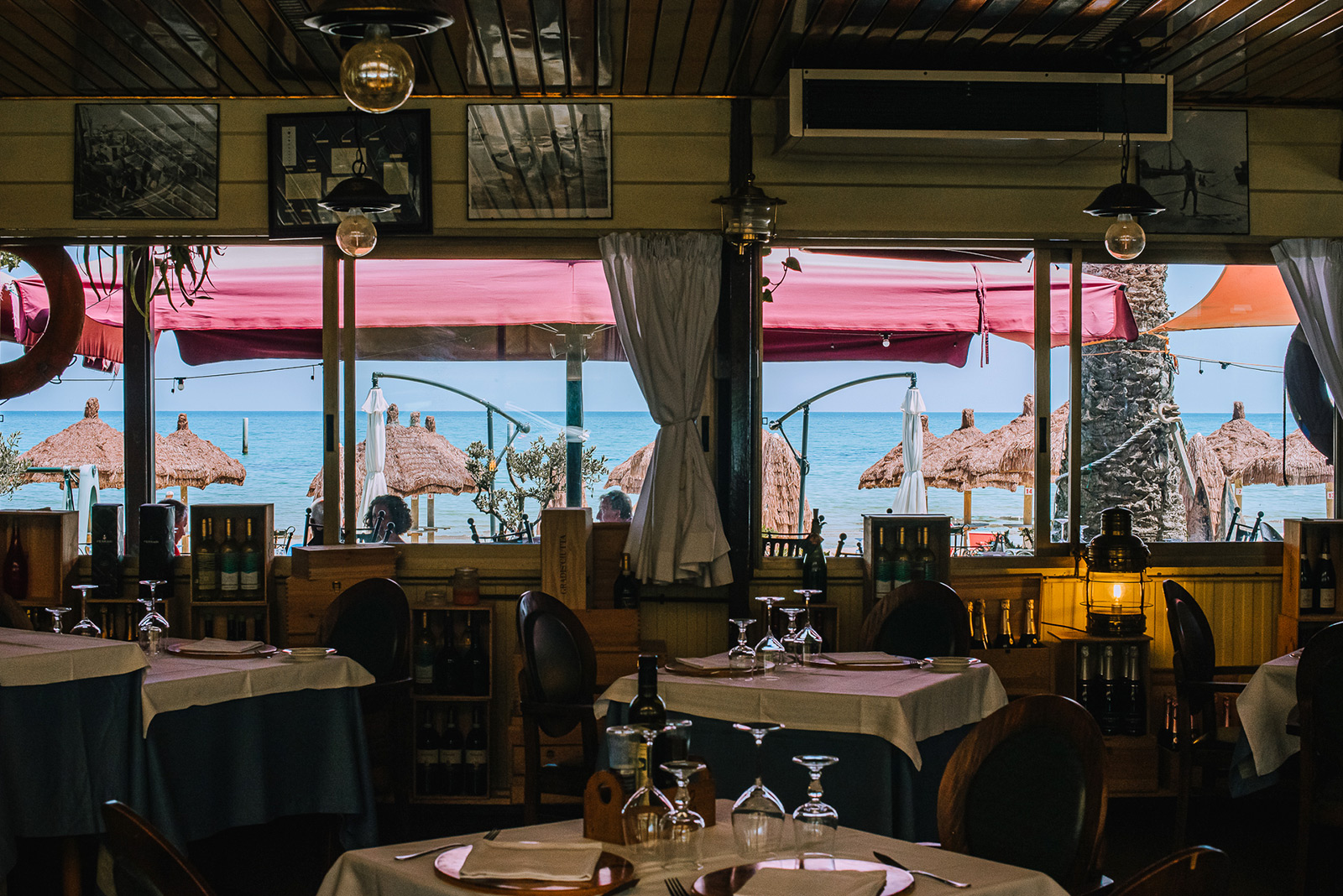 Sala dello Scoglio con tavoli apparecchiati, bottiglie di vino e vetrate su ombrelloni di paglia e mare azzurro