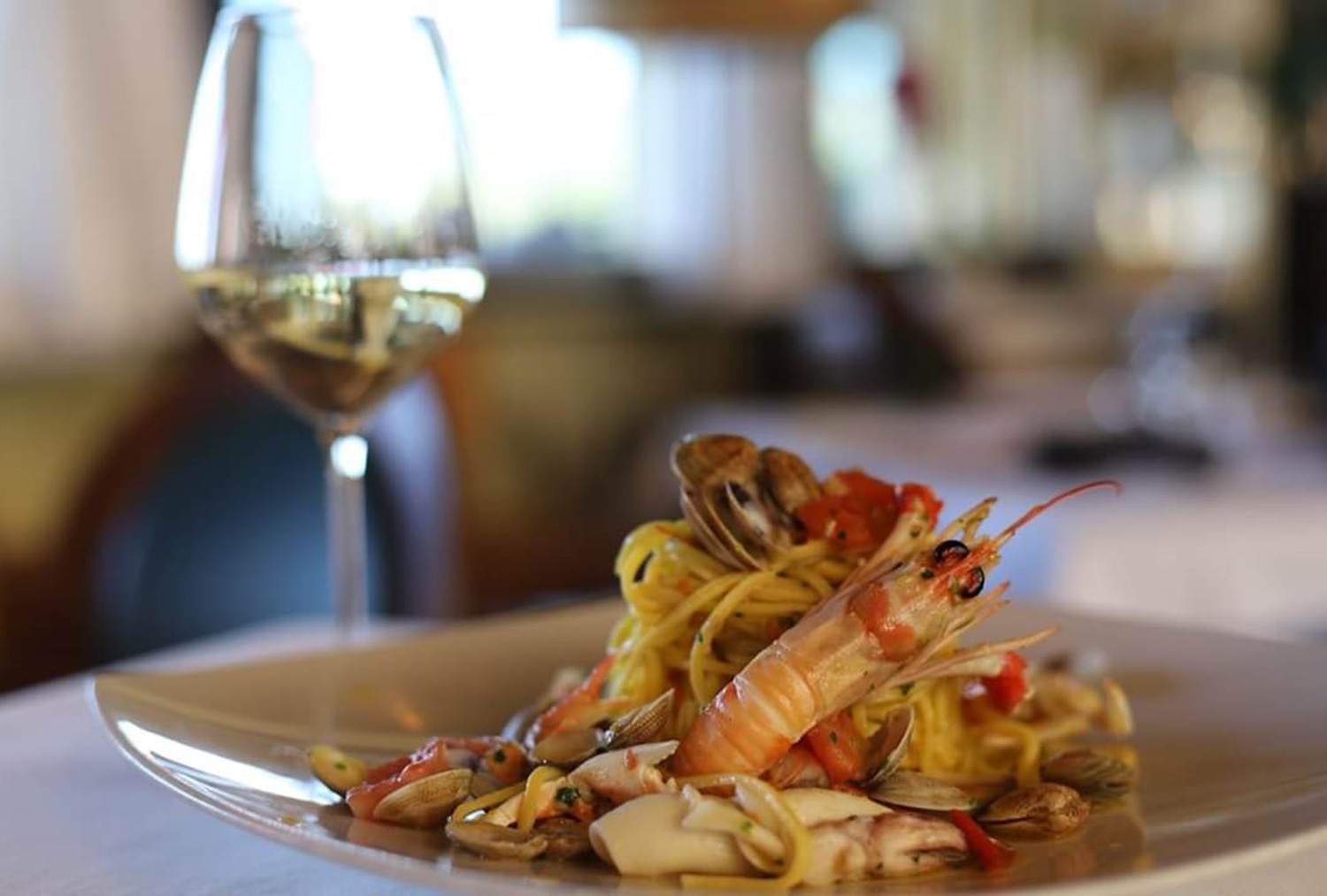 Spaghetti con scampo, vongole, totano e pomodorini in un piatto bianco, accanto a un calice di vino bianco de Lo Scoglio